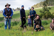 A group of people, including adults and children, standing and kneeling in a grassy field, with a dog at their feet, all participating in land restoration work.